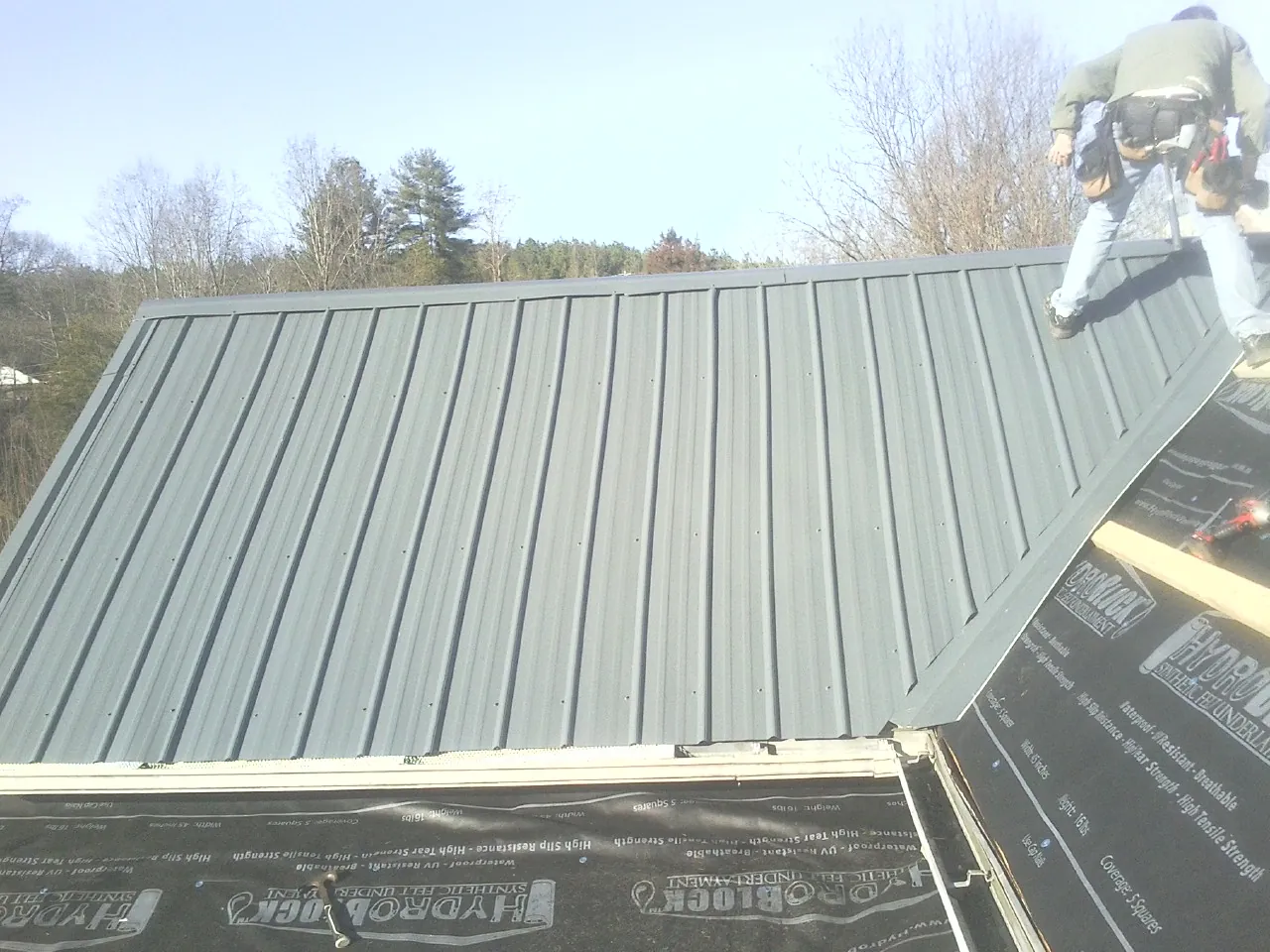 Crew installing dark green standing seam panels over synthetic underlayment in Farley